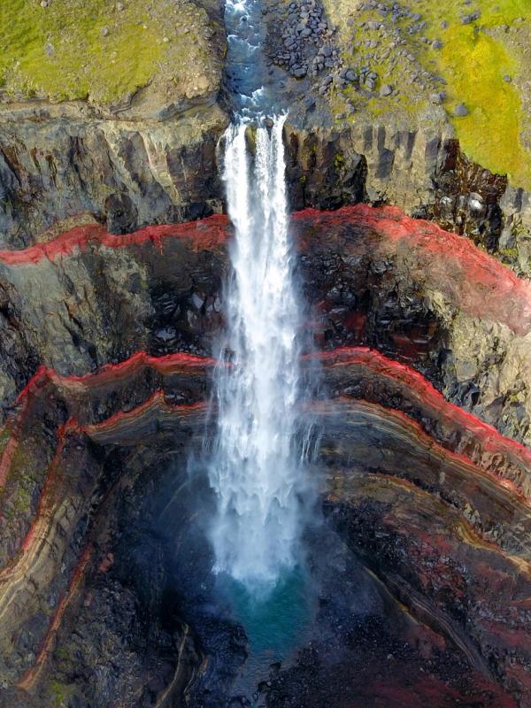 A waterfall cascades down a cliff face revealing prominent red and multicolored geological layers.