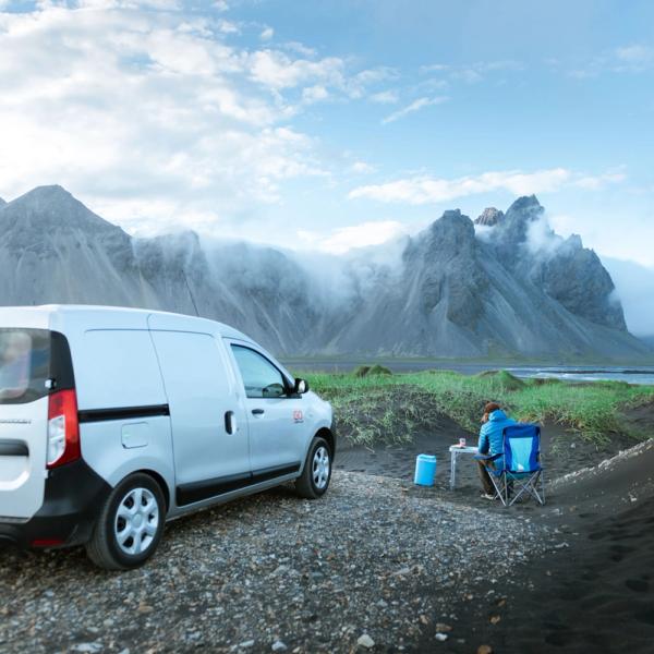 white campervan parked on a black sad beach and a person sitting on a foldable chair outside
