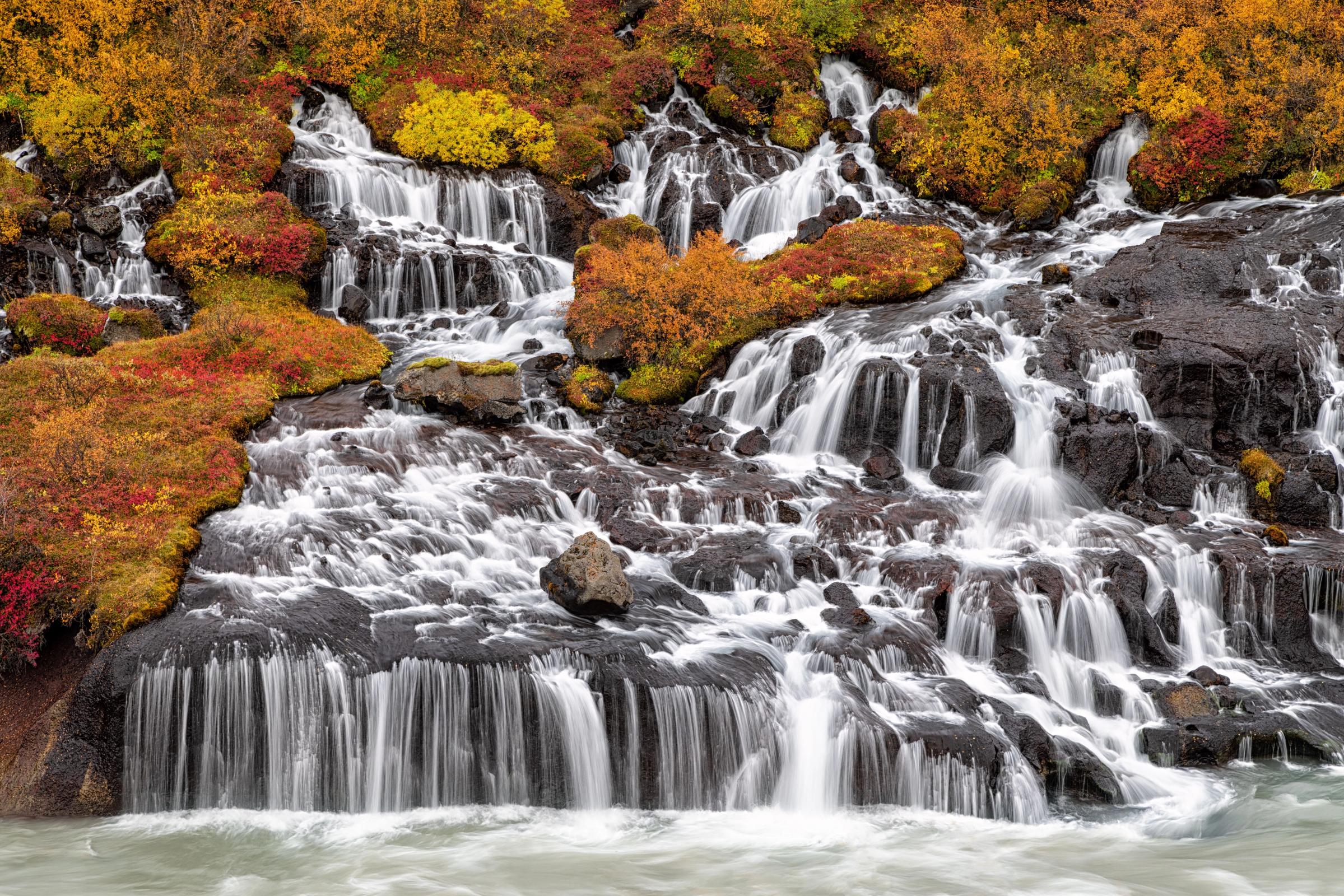 Water cascades over dark, multi-tiered rocks, surrounded by vibrant red, orange, and yellow autumn foliage.