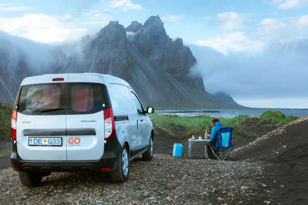 A silver Go Campers van parked on black sand, with a person seated at a table enjoying the view of mist-shrouded mountains and the ocean.