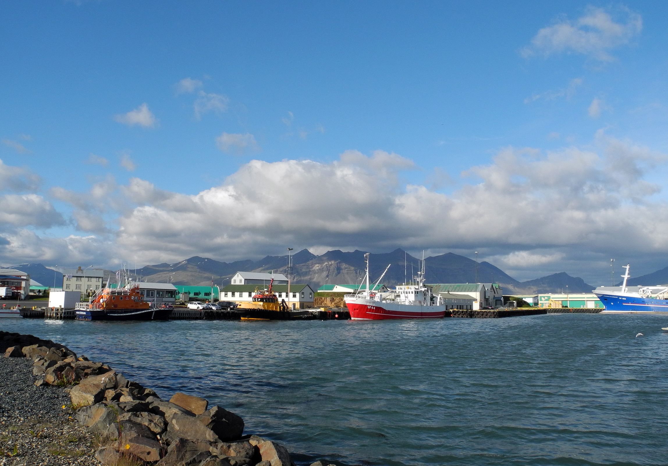 A harbor with colorful boats docked, waterfront buildings, and mountains under a blue, cloudy sky.