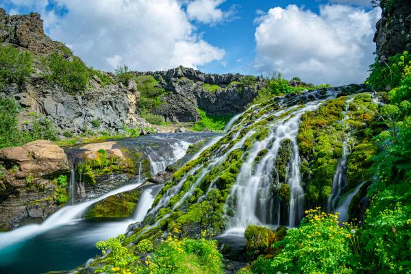 A waterfall flows over bright green mossy rocks into a river, surrounded by dark cliffs and lush plants under a blue, cloudy sky.