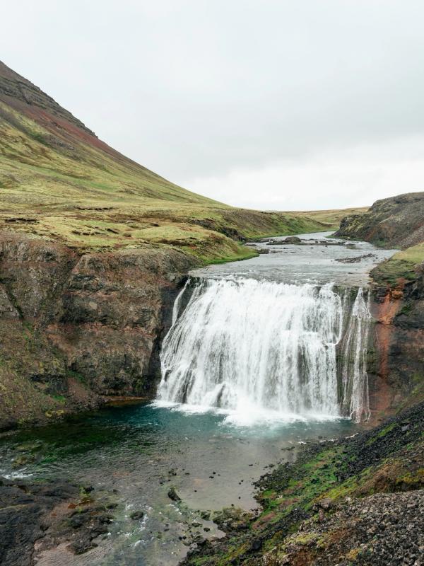 A wide waterfall plunges into a river, flanked by dark, rocky cliffs and green, grassy hills under an overcast sky.