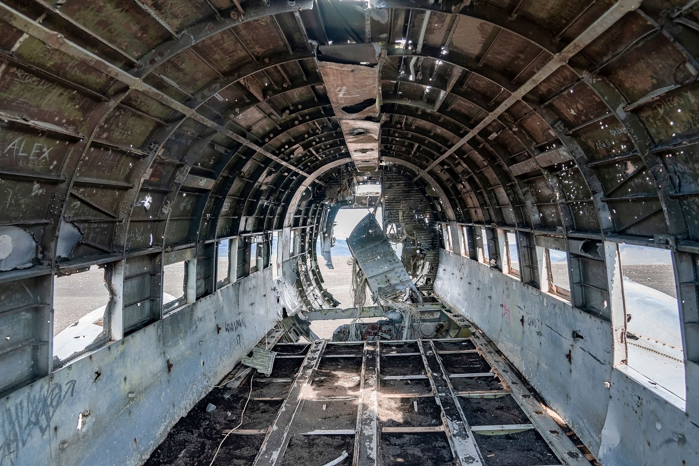 Interior view of a derelict airplane fuselage with exposed metal ribs and empty window frames.
