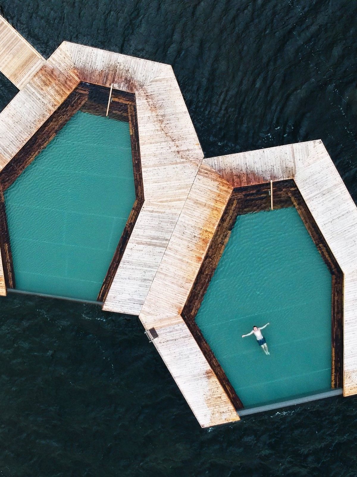 Aerial view of two hexagonal pools with wooden decks on dark water, a person floats in one.
