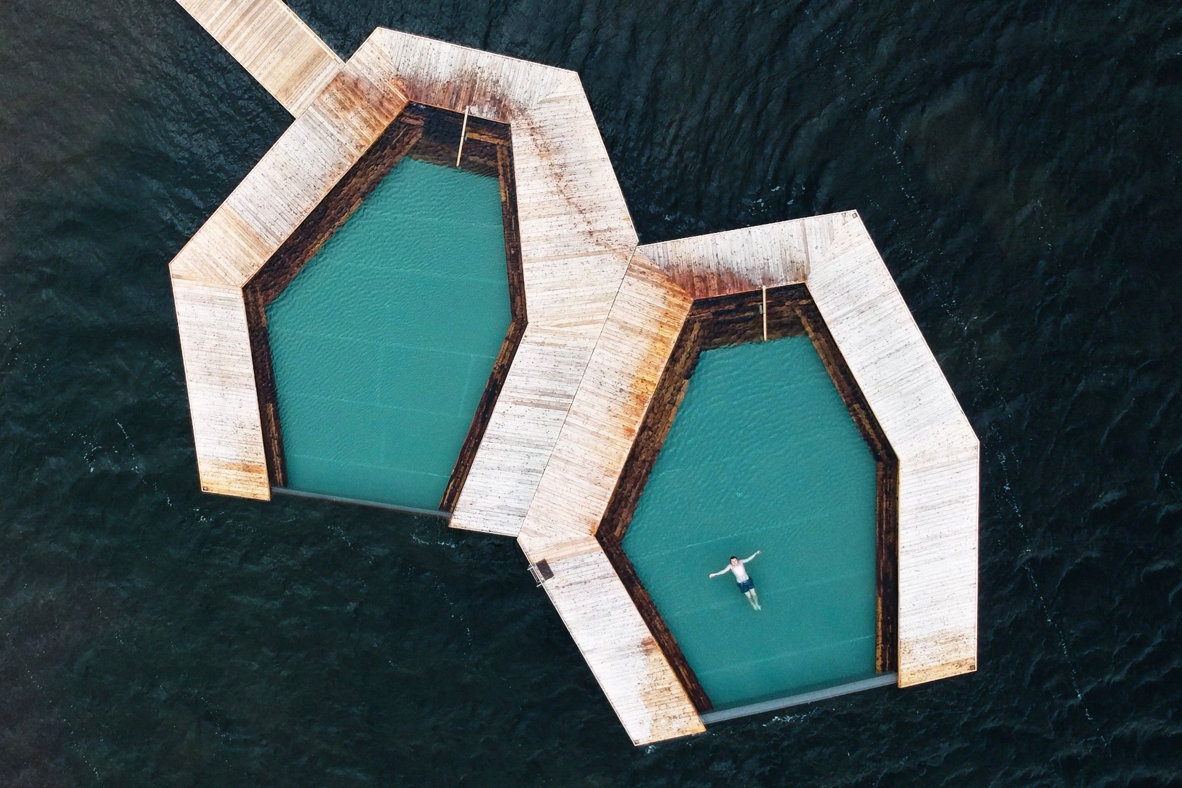 Two hexagonal swimming pools on a wooden platform in dark water, with a person floating in one.