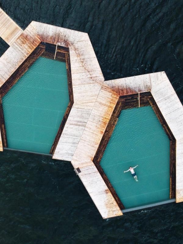 Aerial view of two hexagonal pools with wooden decks on dark water, a person floats in one.