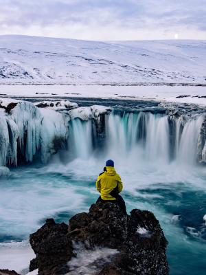 Una persona con una chaqueta amarilla está sentada sobre una roca contemplando una cascada parcialmente congelada en un paisaje nevado.