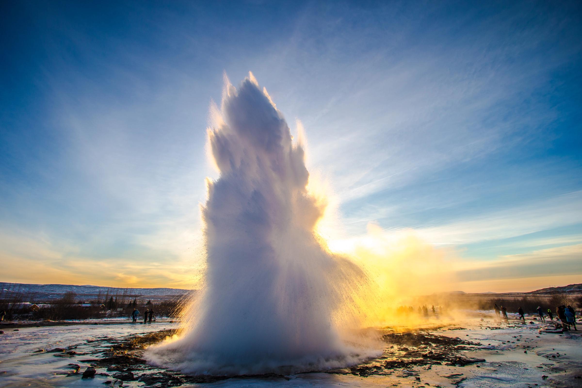 A powerful geyser erupts, shooting a tall column of water and steam high into the air, backlit by golden sunlight.