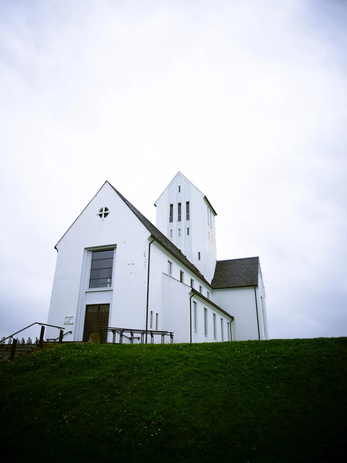 A white church with a tall tower on a grassy hill under an overcast sky.