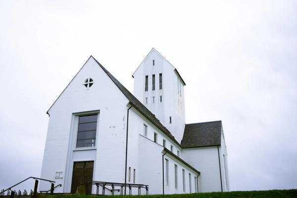 A white church with a tall tower on a grassy hill under an overcast sky.