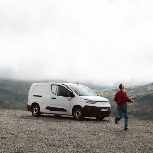 A person runs from a white van on a gravel lot, with misty mountains in the background.