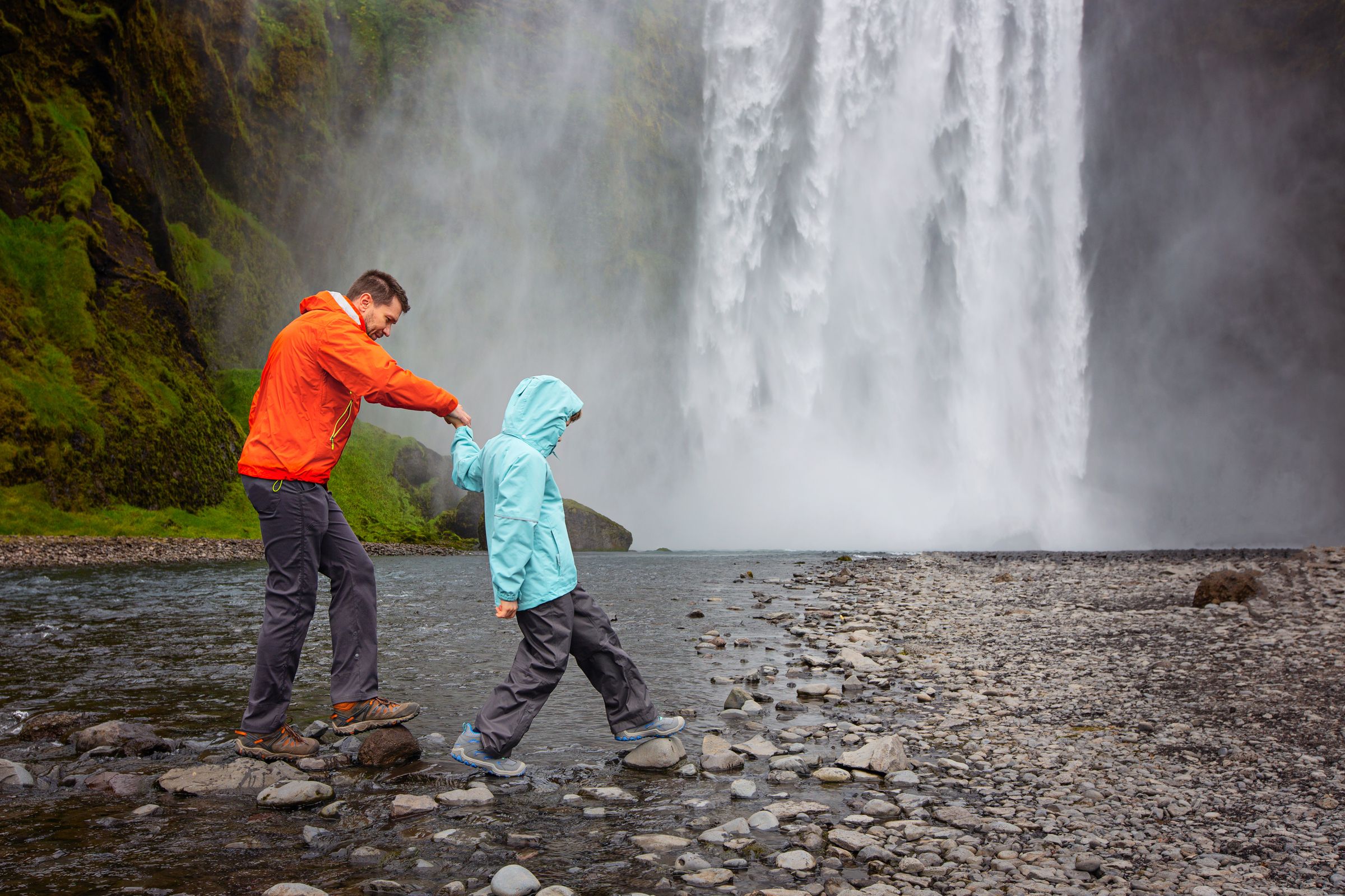 a man and a child are crossing a river near a waterfall in iceland.