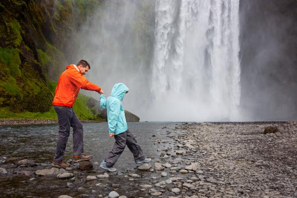 a man and a child are crossing a river near a waterfall in iceland.