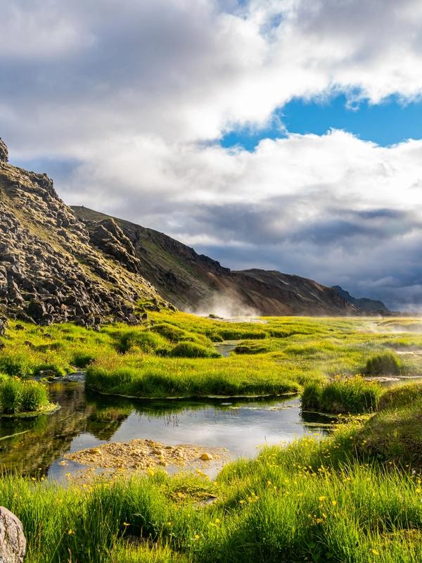 Lush green geothermal field with steam rising from water, flanked by rocky mountains under a cloudy sky.