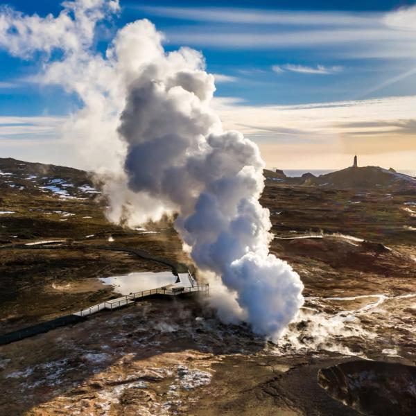 A large plume of steam rises from a geothermal area with a boardwalk in a rocky landscape, a lighthouse visible in the distance under a bright sky.