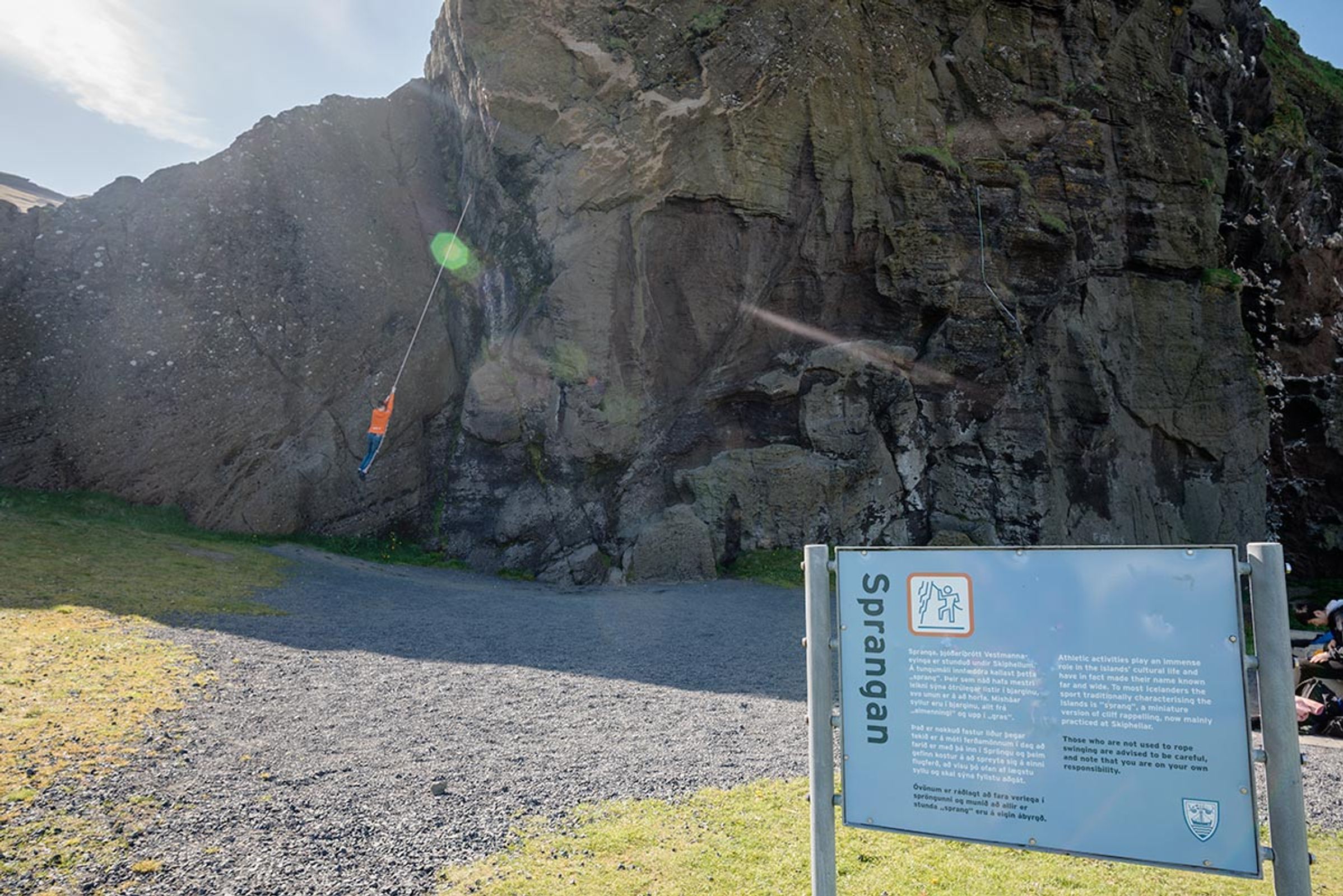 A person hangs by a rope against a large cliff face, with an informational sign about Sprangan in the foreground.