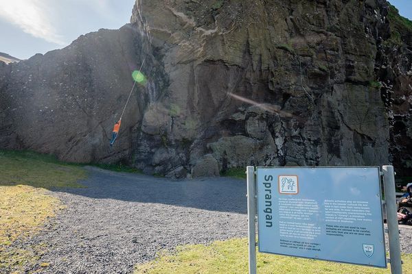 A person hangs by a rope against a large cliff face, with an informational sign about Sprangan in the foreground.