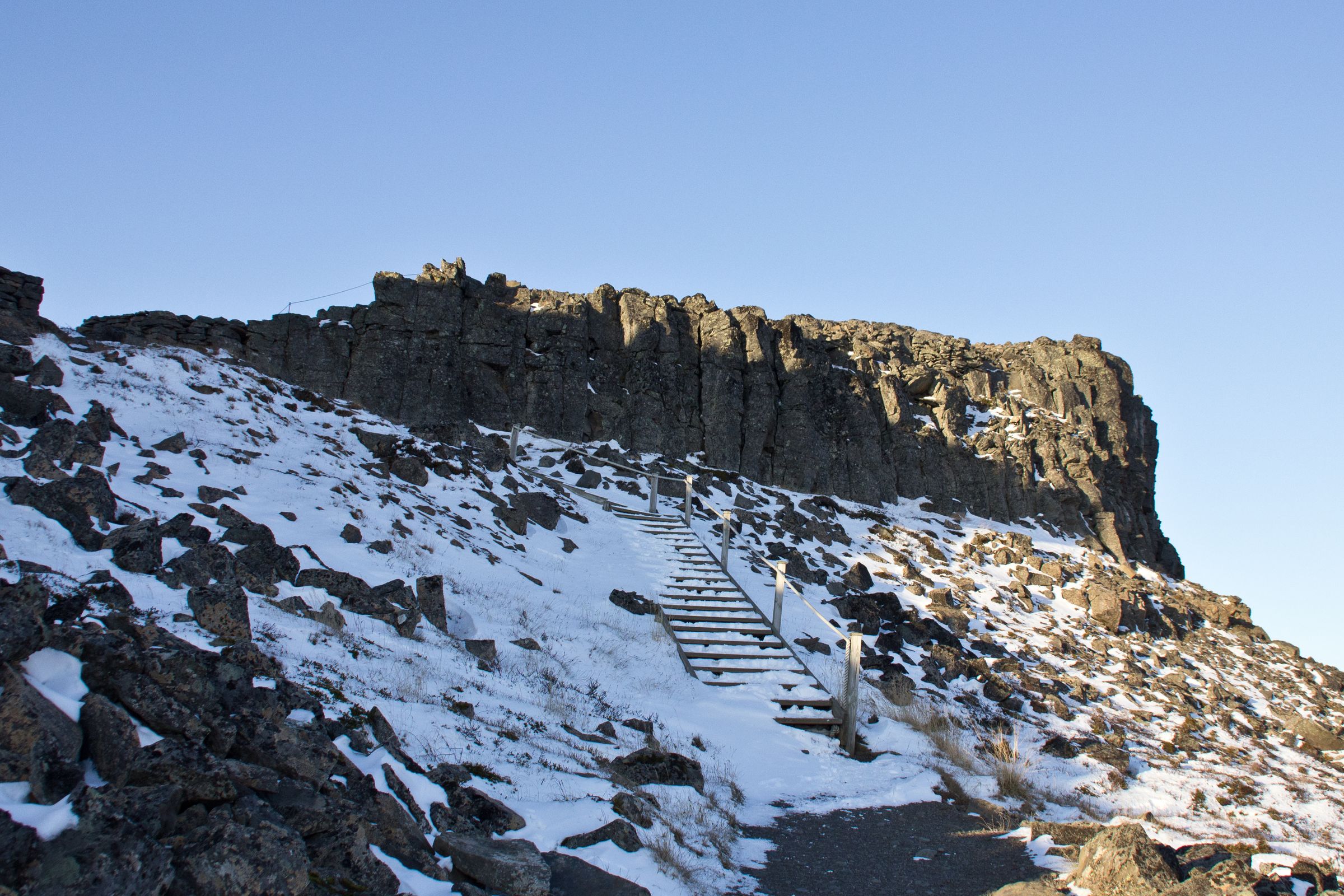 A wooden staircase ascends a rocky, snow-dusted slope towards a dark cliff under a clear blue sky.