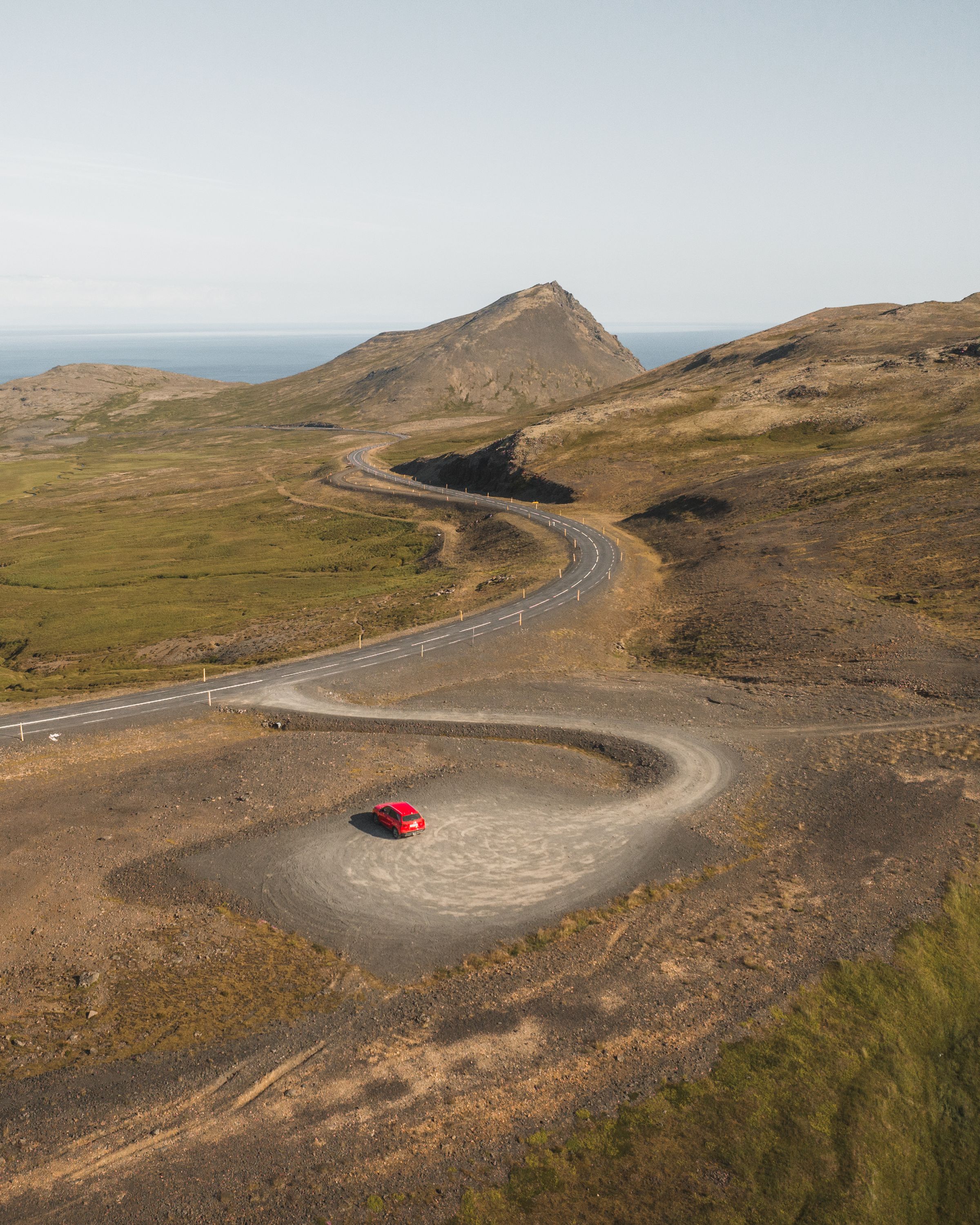 Petite Suzuki Vitara rouge sur une route goudronnée sinueuse, avec le littoral au loin, autotour en Islande