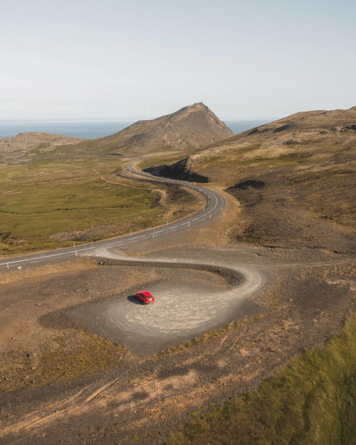 Petite Suzuki Vitara rouge sur une route goudronnée sinueuse, avec le littoral au loin, autotour en Islande
