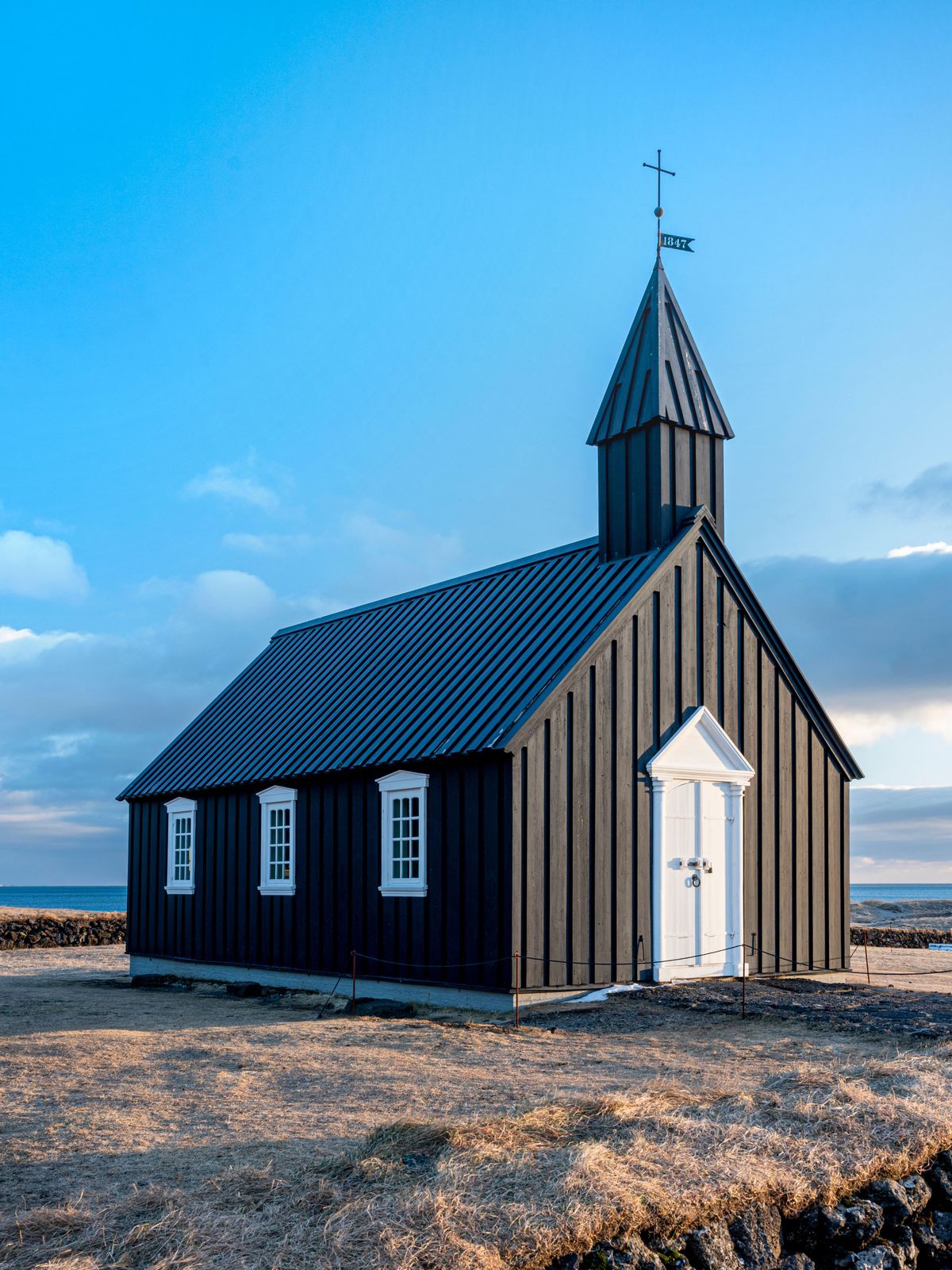 a small black church with a cross on top of it is sitting on top of a hill next to the ocean .