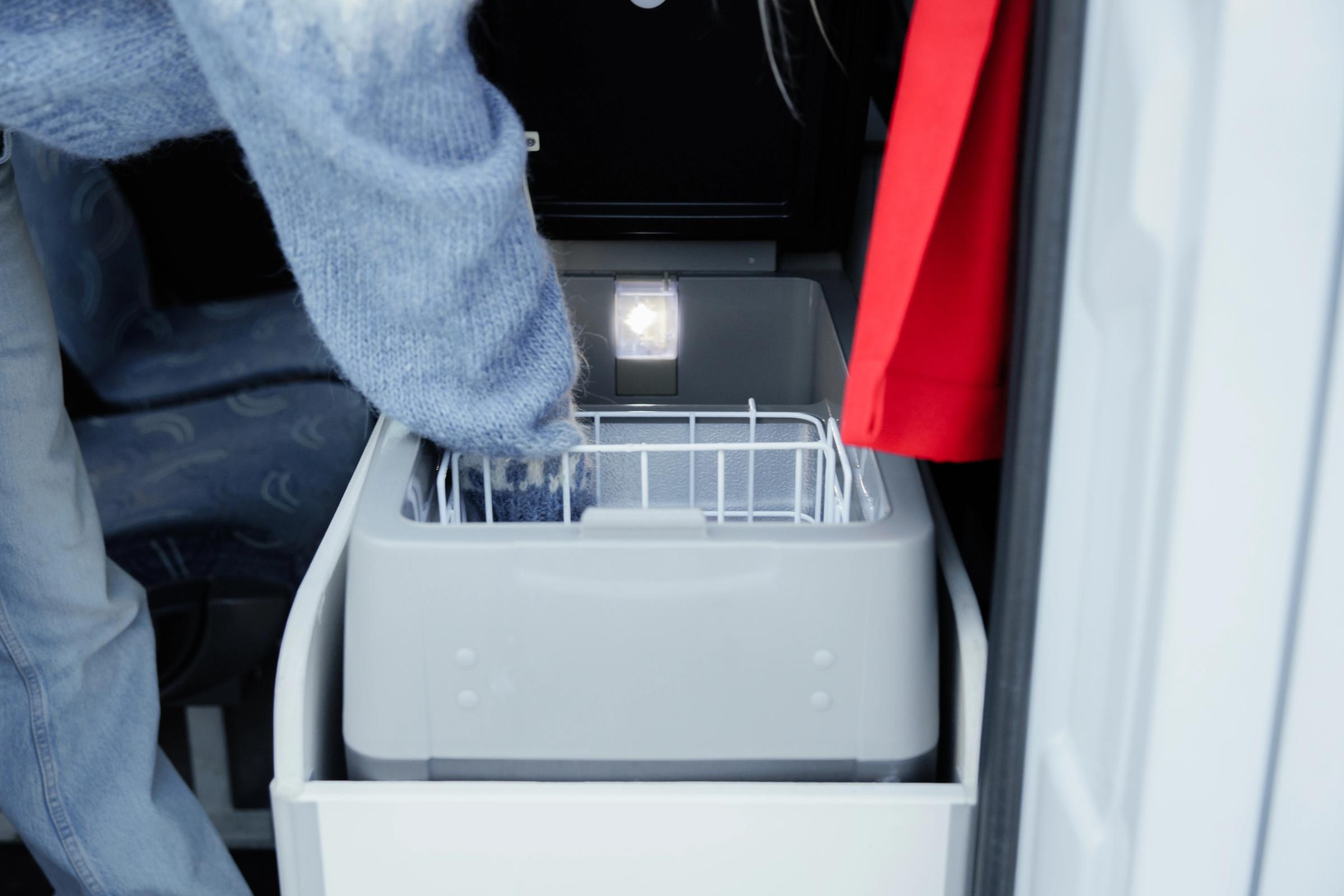 Eine Person greift in eine geöffnete Kühlbox mit eingeschalteter Innenbeleuchtung und einem Drahtkorb im Inneren