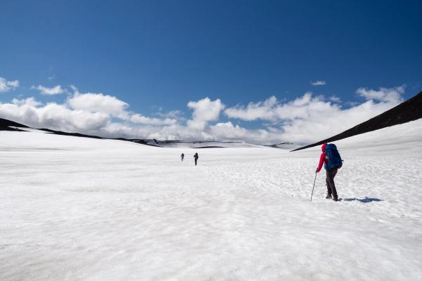 Hikers cross a vast snowfield with dark mountains under a blue, cloudy sky.