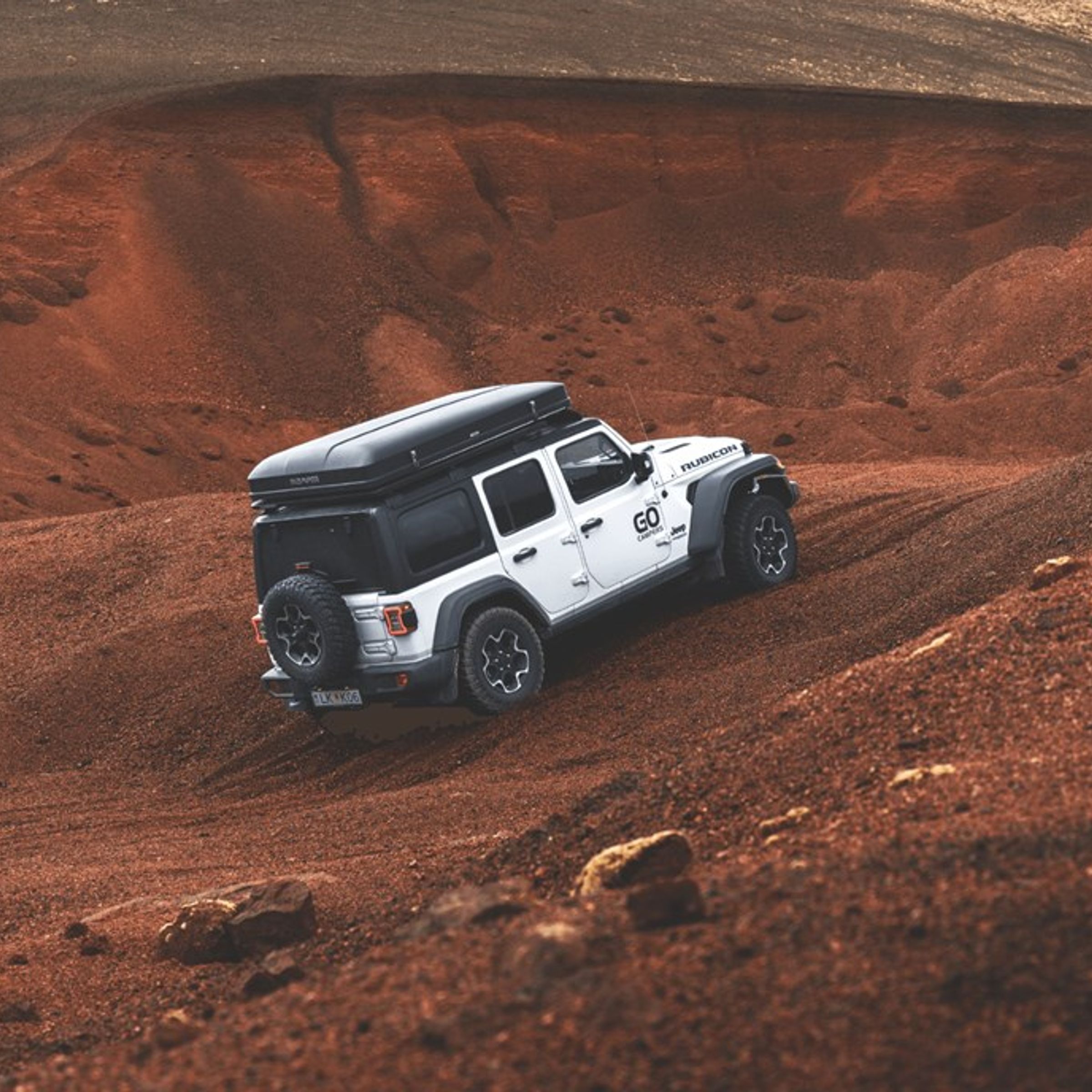 White Jeep Wrangler with a roof-top tent climbing a steep, red dirt hill.