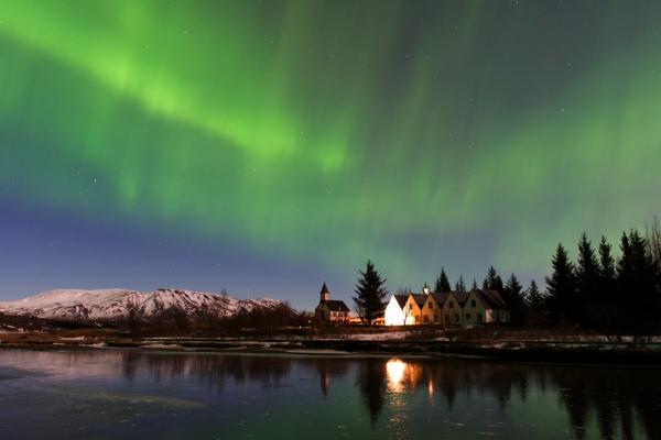 La Aurora Boreal sobre unas casas iluminadas junto a un bosque y un lago