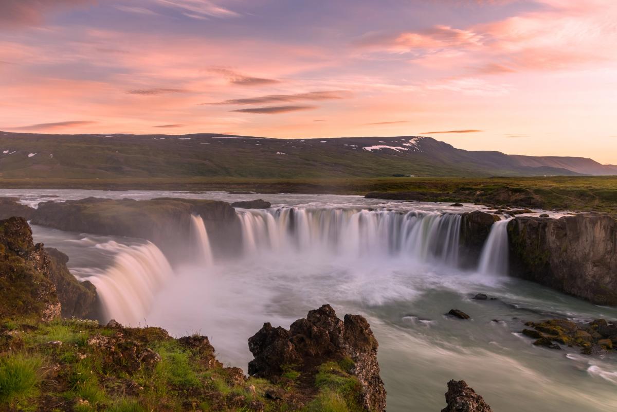 a waterfall in the middle of a river with mountains in the background at midnight sun in iceland .