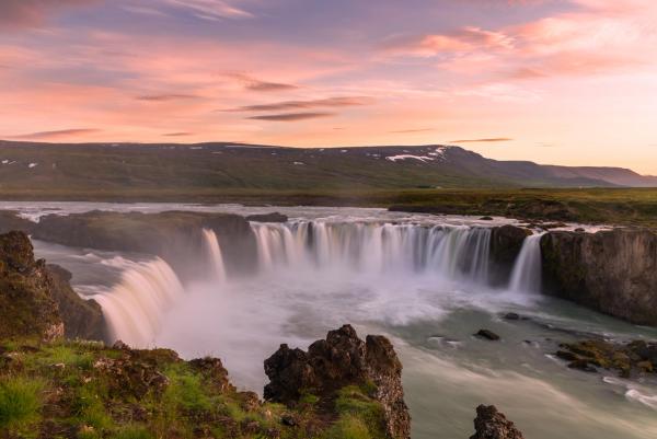 a waterfall in the middle of a river with mountains in the background at midnight sun in iceland .