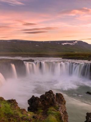 a waterfall in the middle of a river with mountains in the background at midnight sun in iceland .