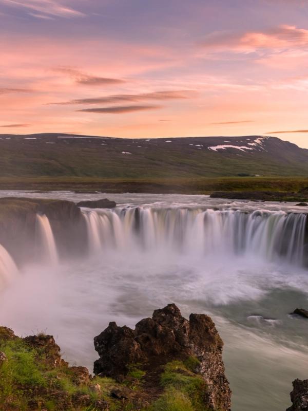 a waterfall in the middle of a river with mountains in the background at midnight sun in iceland .