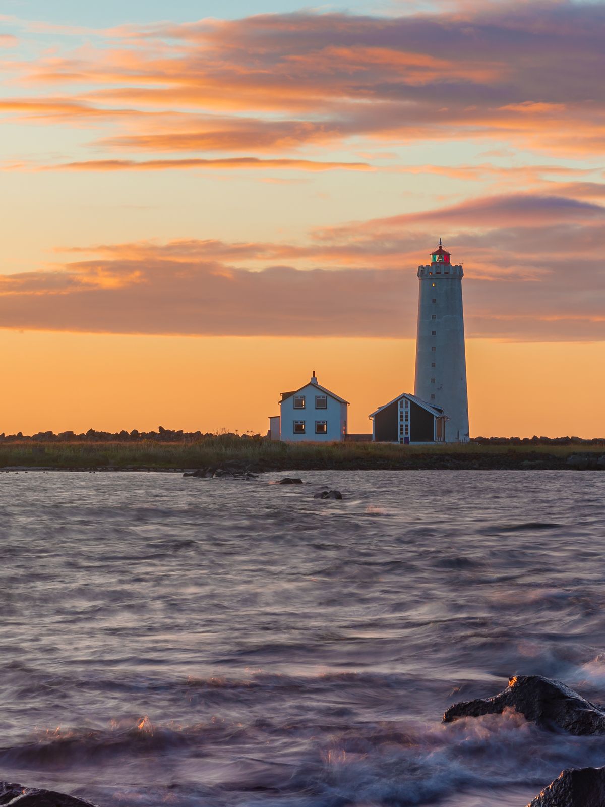 A white lighthouse near the sea with two houses next to it during sunset