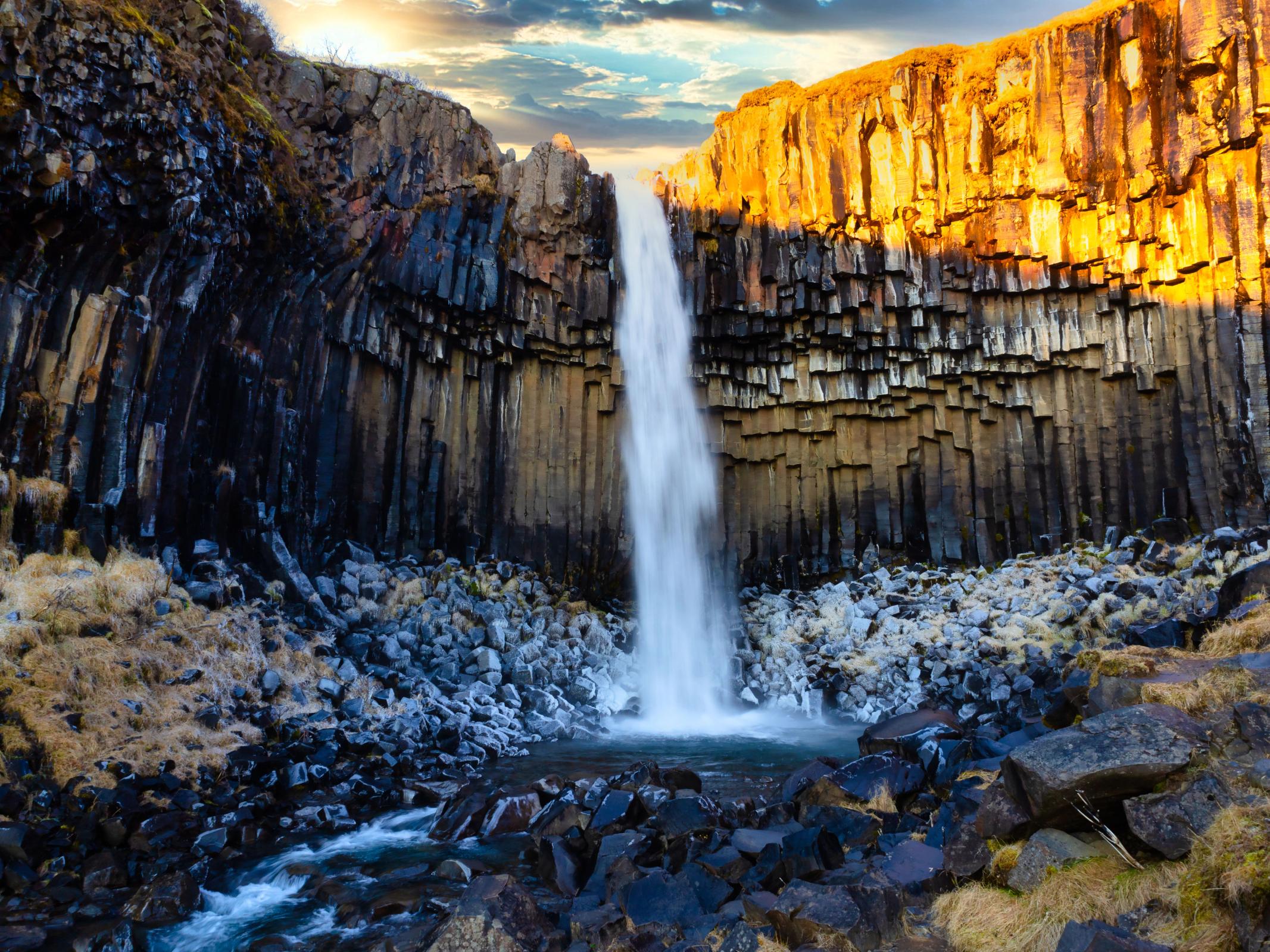 Waterfall cascading over dark hexagonal basalt columns, with one side brightly lit by warm sunlight.