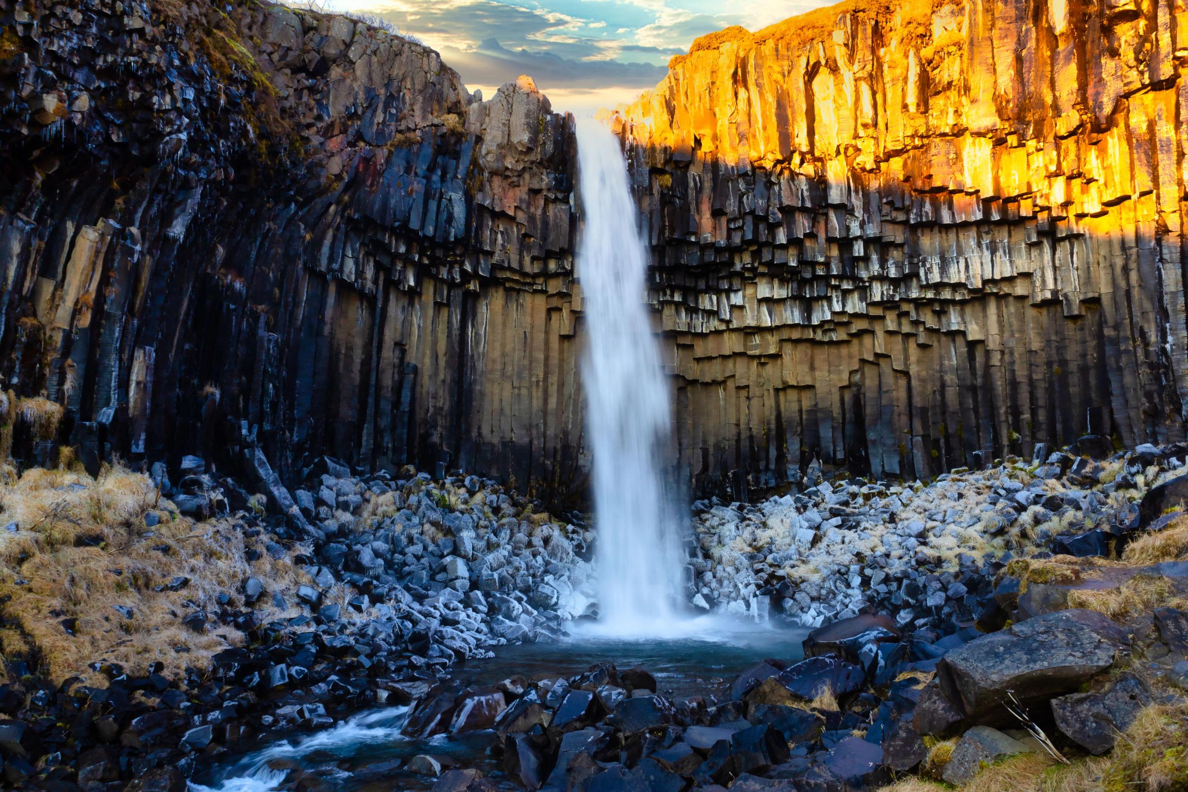 Waterfall cascading over dark hexagonal basalt columns, with one side brightly lit by warm sunlight.