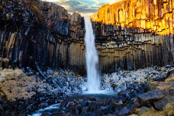 Waterfall cascading over dark hexagonal basalt columns, with one side brightly lit by warm sunlight.