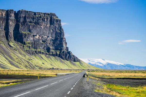 Una carretera se extiende junto a un enorme acantilado y campos verdes hacia unas lejanas montañas cubiertas de nieve.