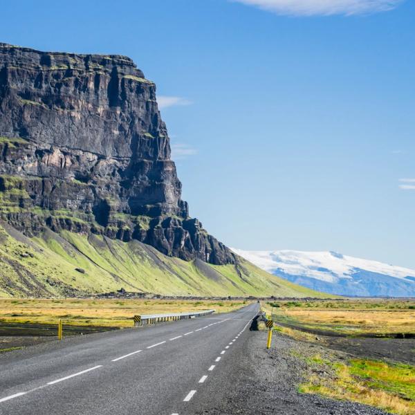 A road extends towards a distant snow-capped peak, flanked by a massive dark cliff on the left and open plains on the right.