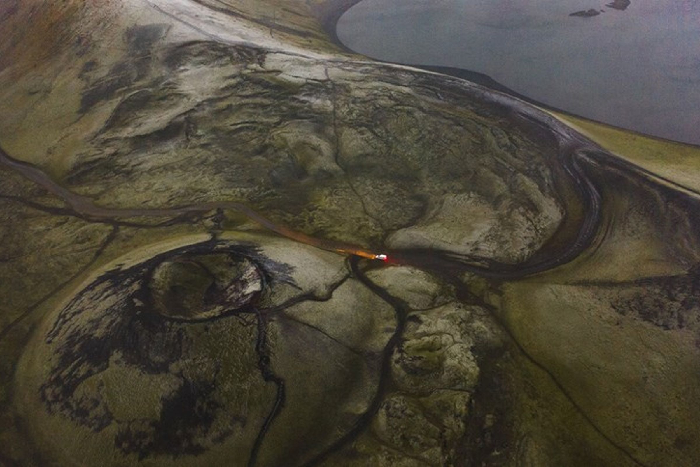 Aerial view of a desolate, muted landscape with a red vehicle driving on a winding path, a small crater, and a body of water.