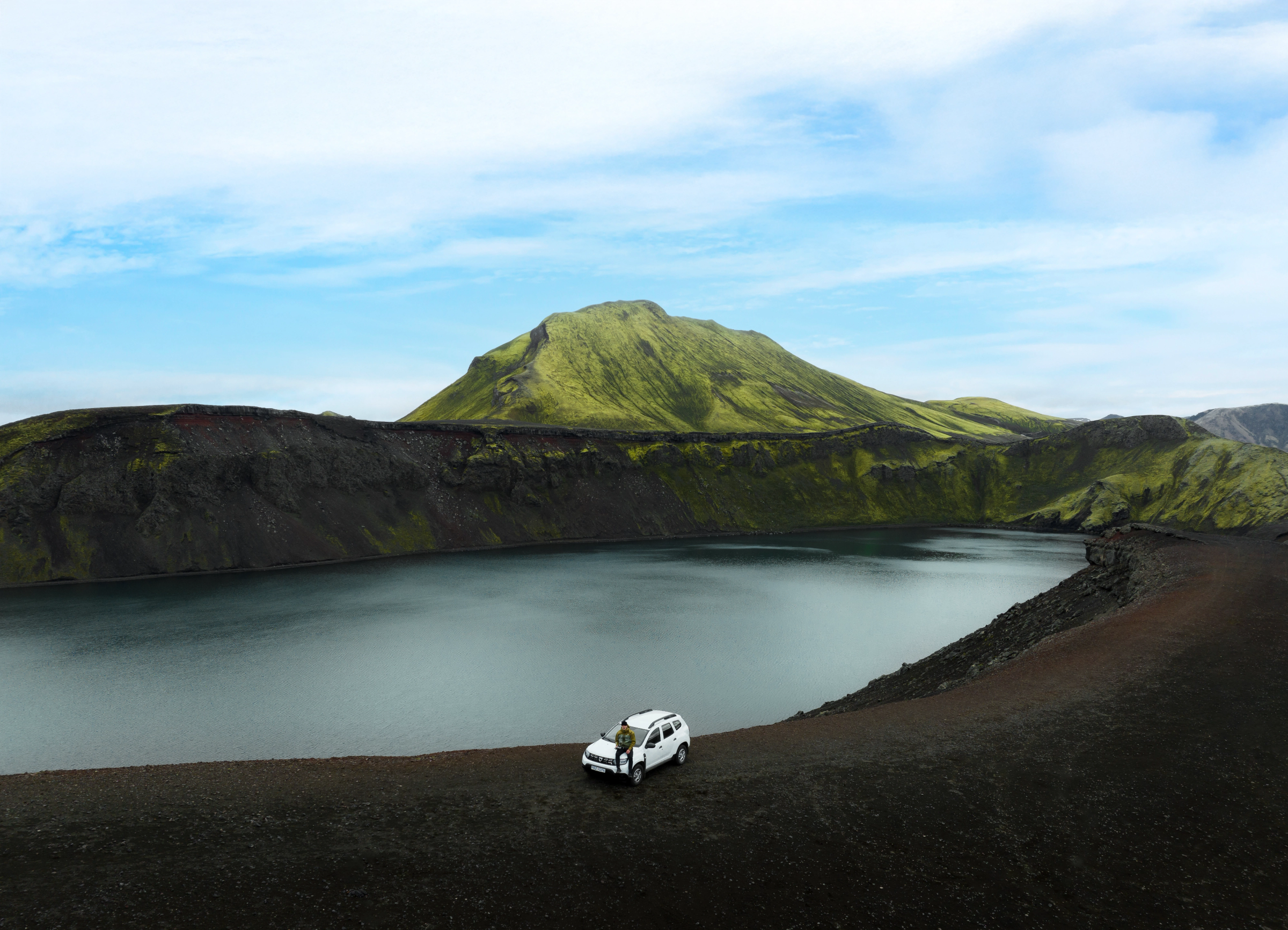 A person stands by a white SUV on dark volcanic ground next to a calm lake, with green mossy mountains in the background.