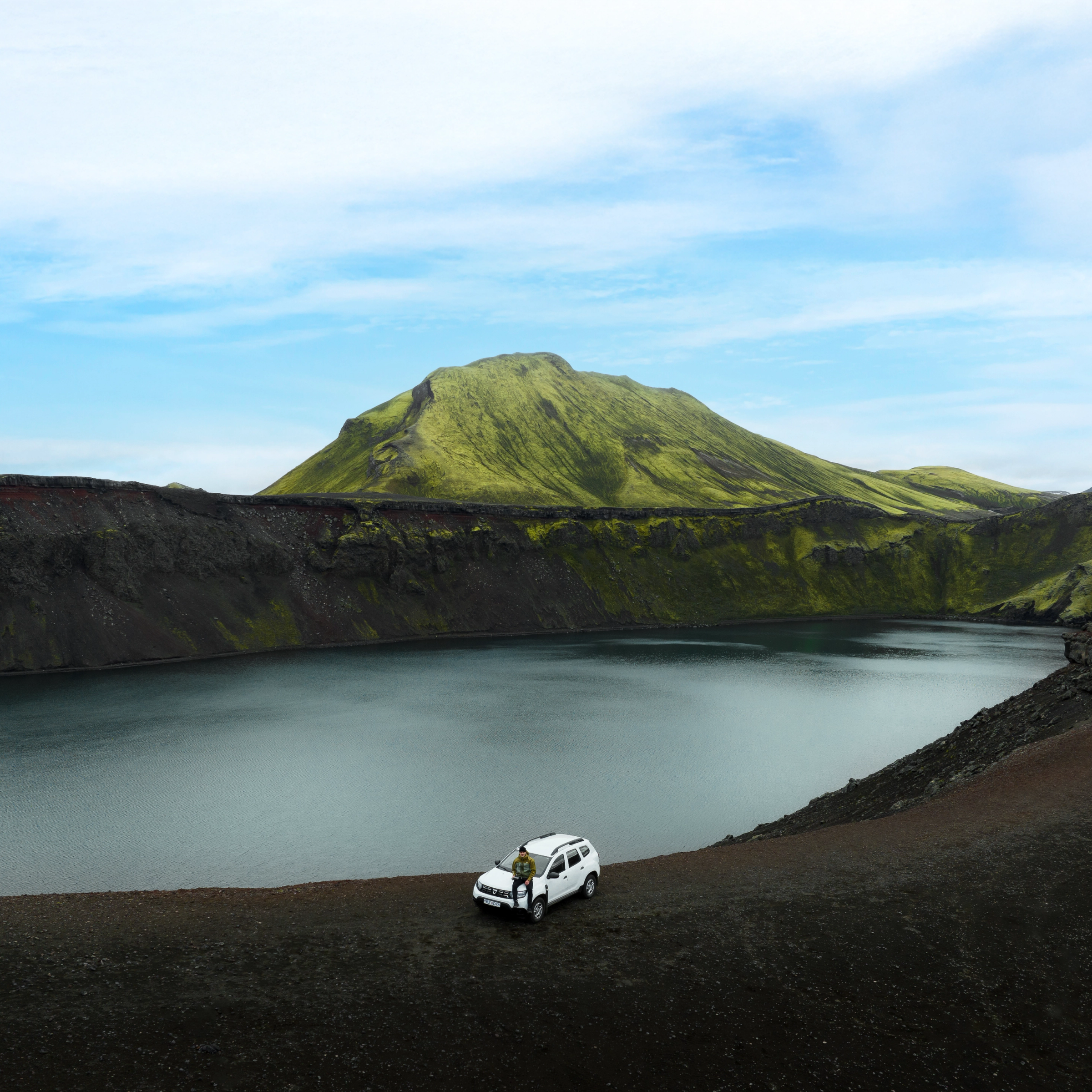 Una persona está junto a un SUV blanco sobre un terreno volcánico oscuro, al lado de un lago tranquilo, con montañas verdes cubiertas de musgo al fondo.