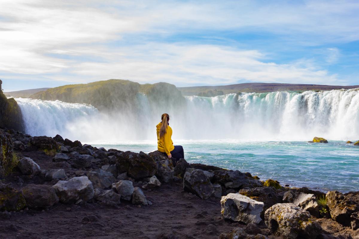 a woman is sitting on a rock near a waterfall taking a picture at Goðafoss in iceland.
