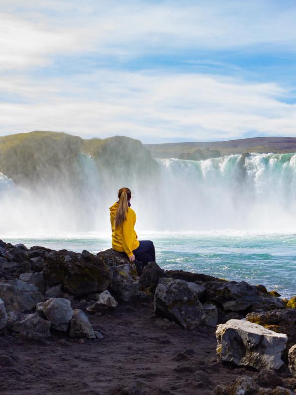 a woman is sitting on a rock near a waterfall taking a picture at Goðafoss in iceland.