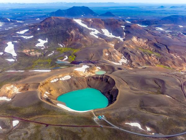 Aerial view of a volcanic landscape featuring a vibrant turquoise crater lake, patches of snow, and colorful barren terrain.