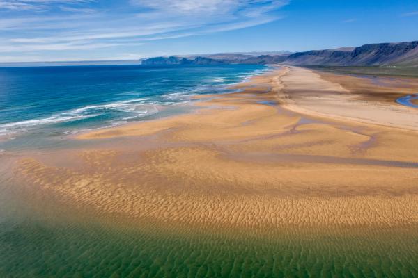 Aerial view of a vast golden sand beach with blue ocean, green shallows, and distant cliffs under a blue sky.
