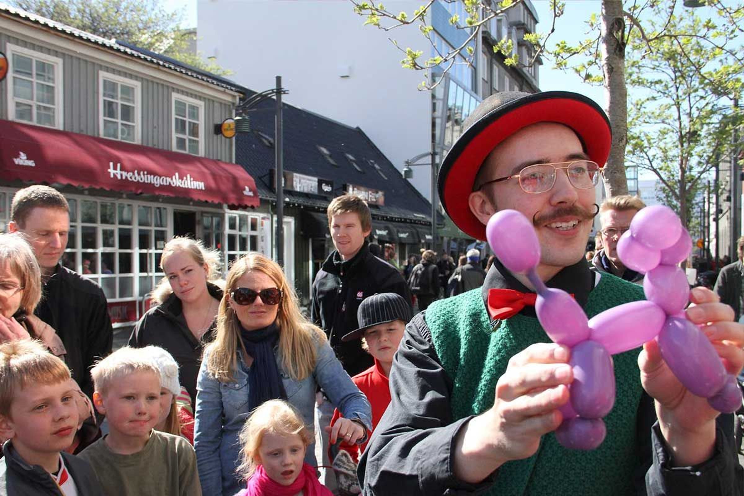 A street performer with a mustache, top hat, and bow tie holds a purple balloon poodle for an audience of children.