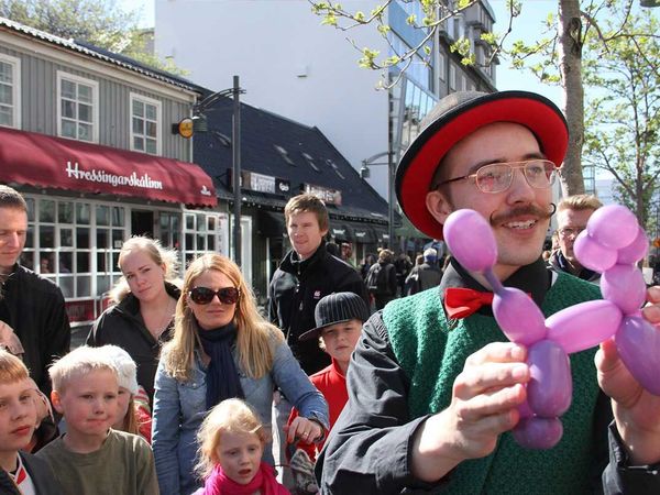 A street performer with a mustache, top hat, and bow tie holds a purple balloon poodle for an audience of children.