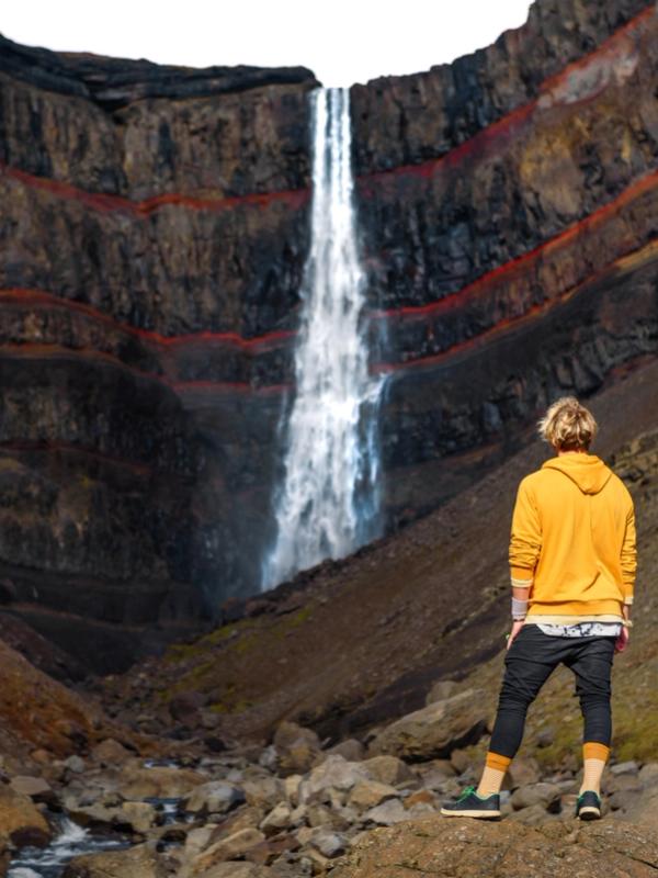 A person in a yellow hoodie stands on rocks looking at a tall waterfall cascading down a dark, layered cliff with red bands.